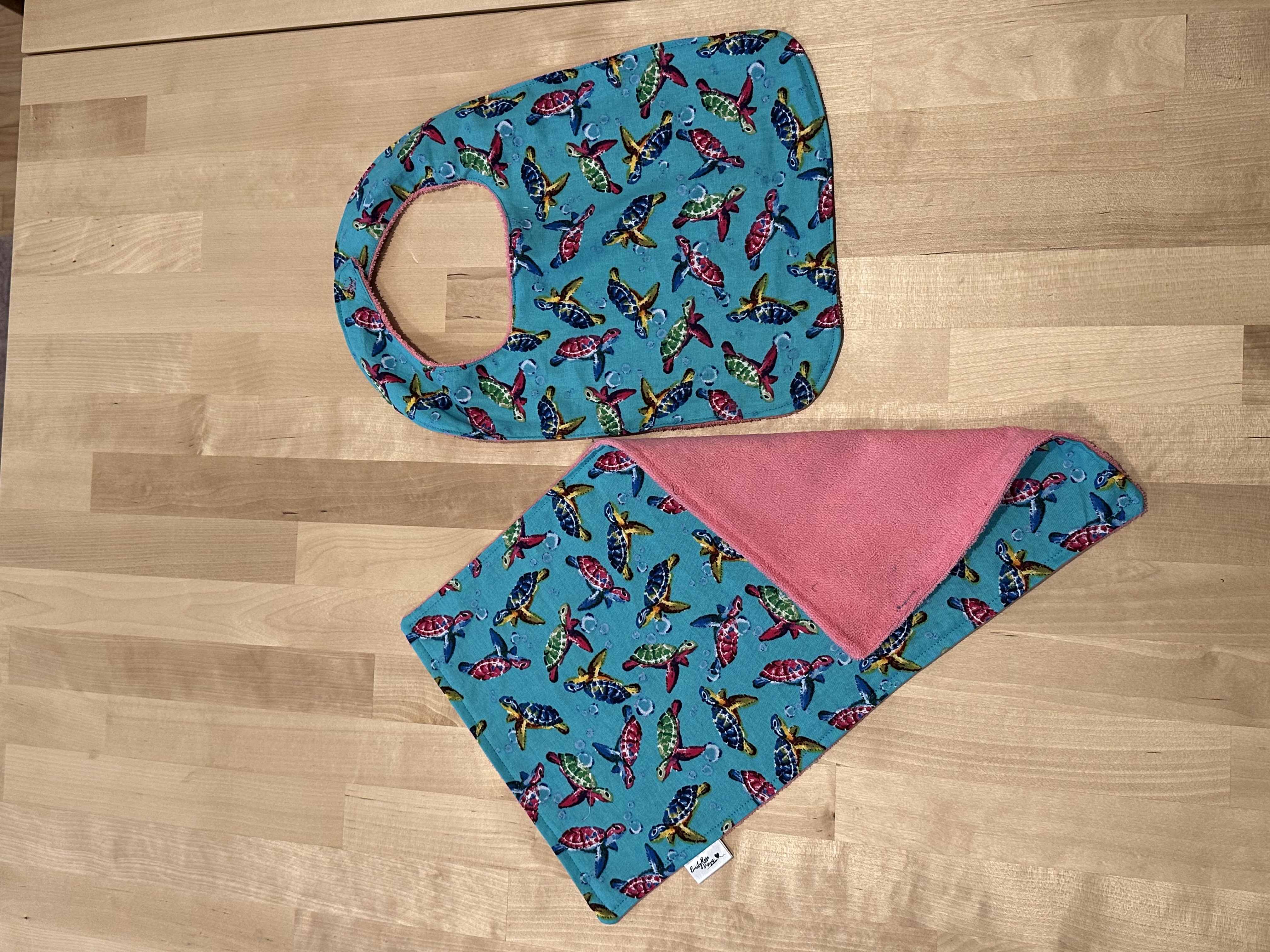 A finished bib and burp cloth set in a colorful sea turtle print on teal, with pink terry on the reverse
