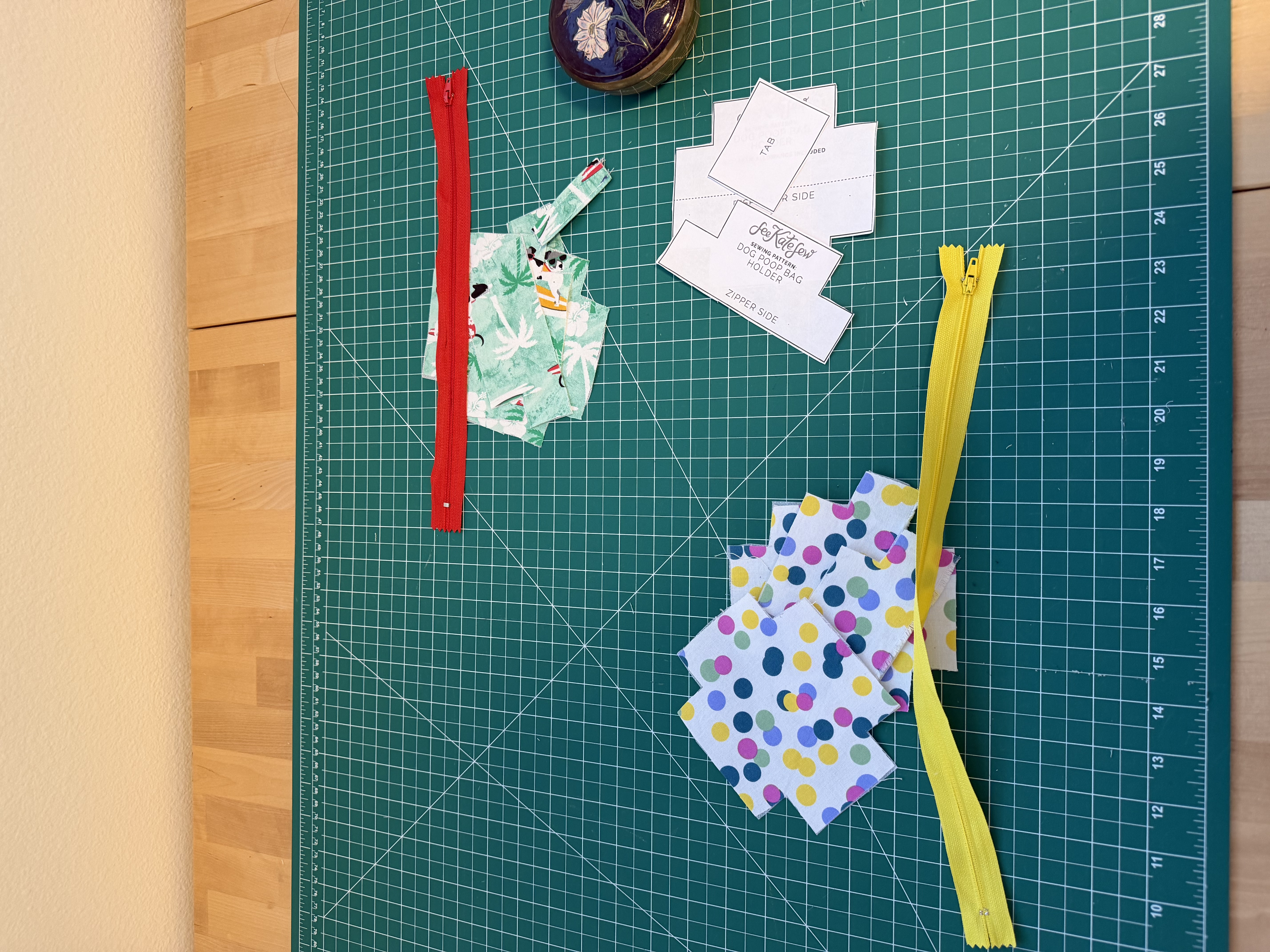 Pattern pieces and cut fabric laid out for two dog poop bag holders, one in a tropical surfing dogs print and one in a colorful polka dot print