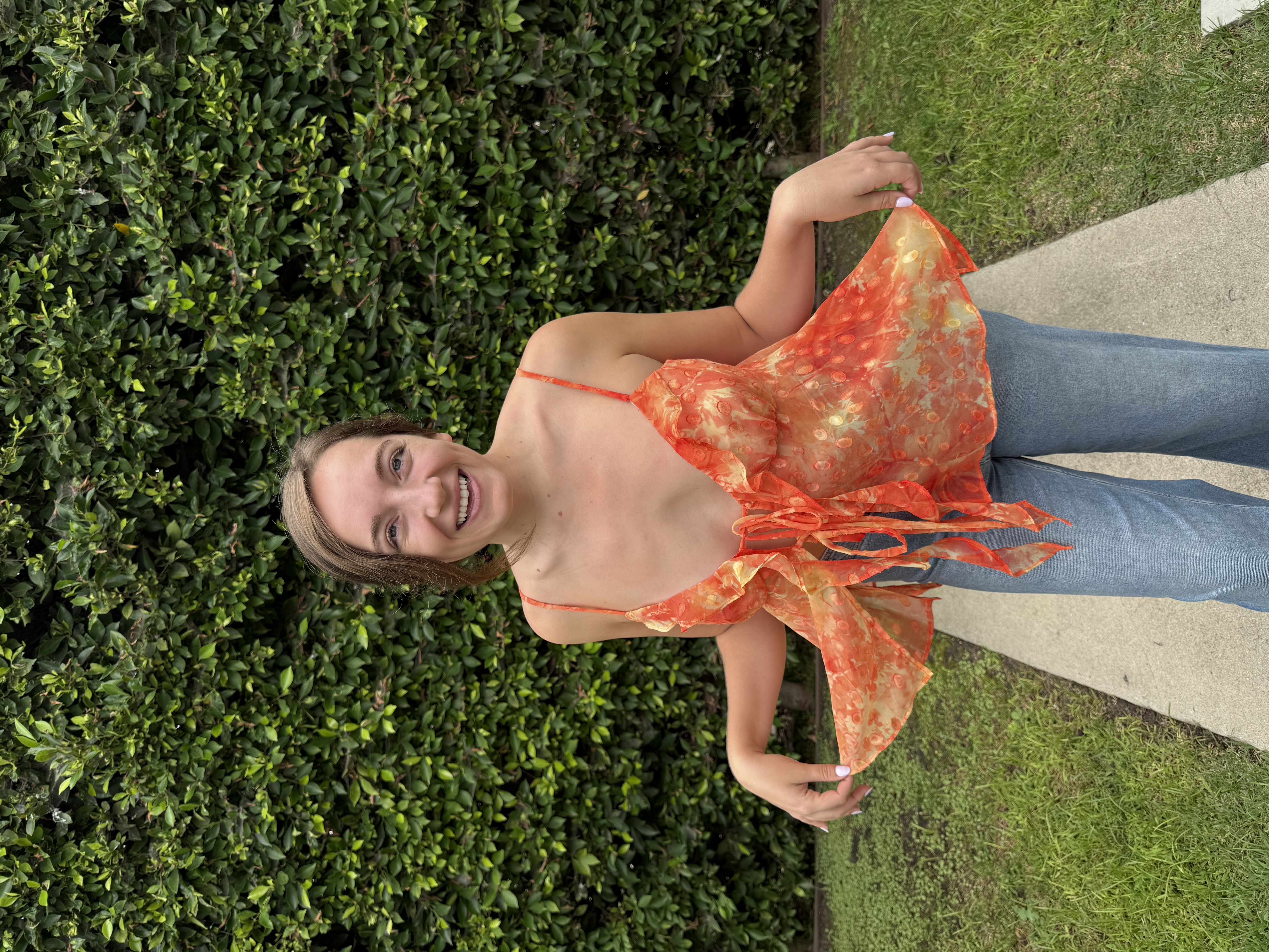 Emily holding out the flounce of the finished Jaya Top in front of a green hedge